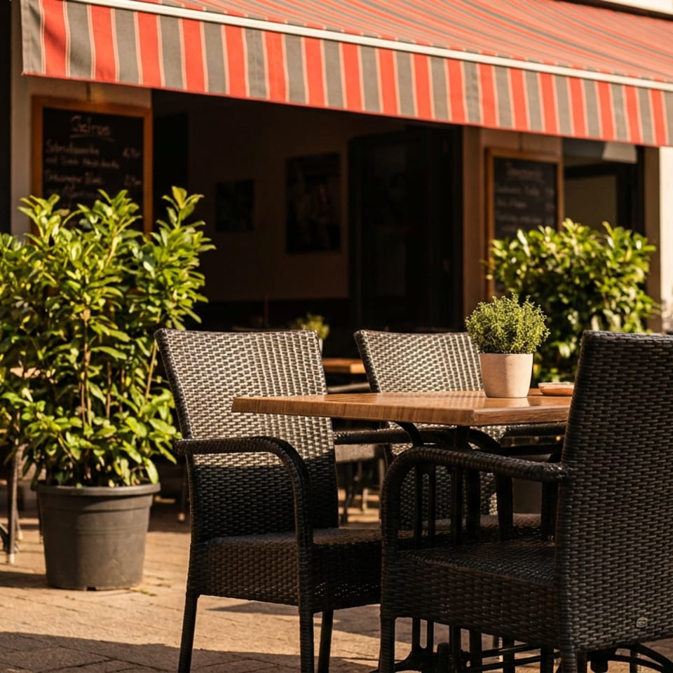 Sunny outdoor restaurant patio with wicker chairs, wooden tables, potted plants, and a red striped awning.
