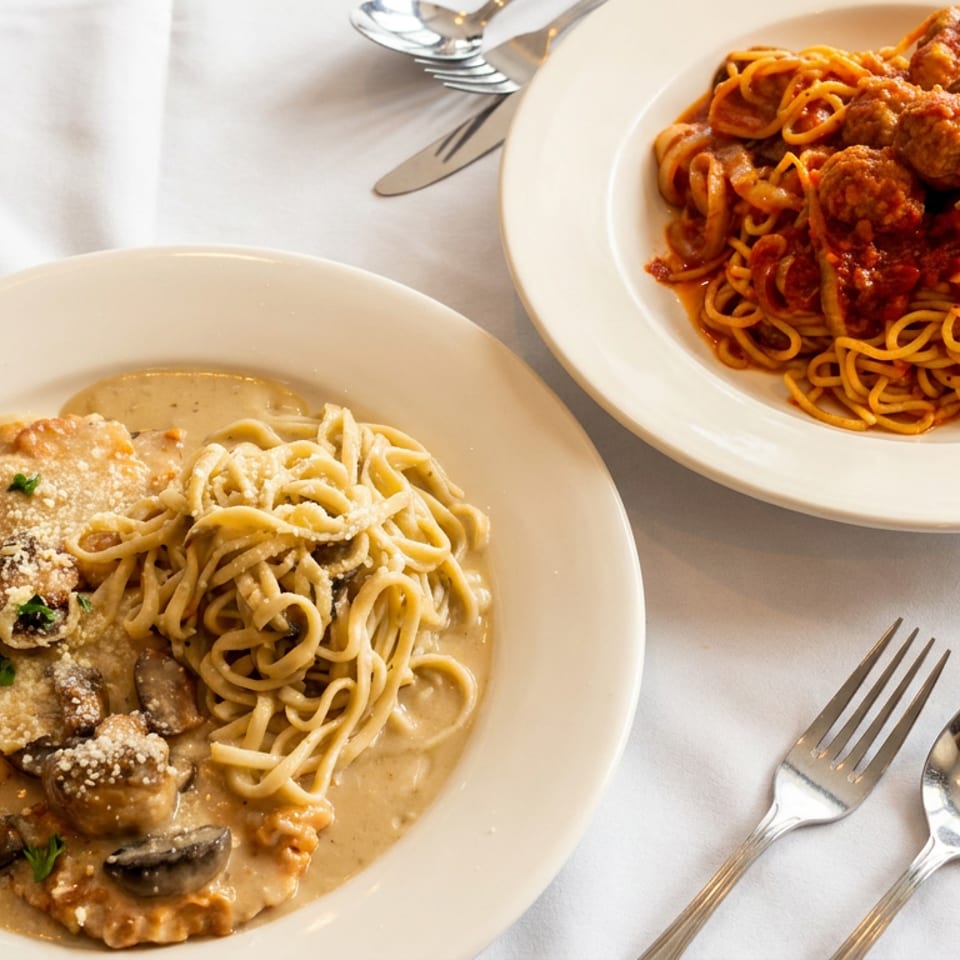 Two pasta dishes: spaghetti with meatballs and chicken with creamy mushroom sauce on a white table.